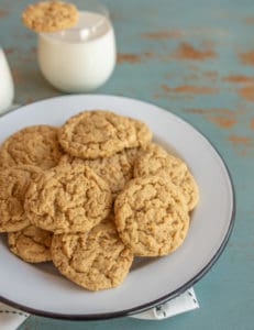 A white plate piled with peanut butter oatmeal cookies sits on a blue rustic table. In the background, two glasses of milk are visible, one with a cookie resting on top.