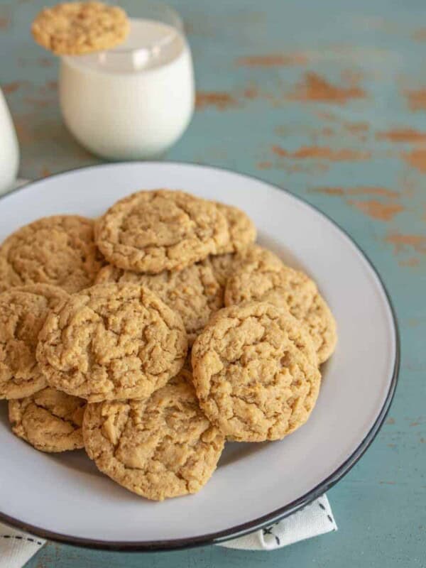 A white plate piled with peanut butter oatmeal cookies sits on a blue rustic table. In the background, two glasses of milk are visible, one with a cookie resting on top.