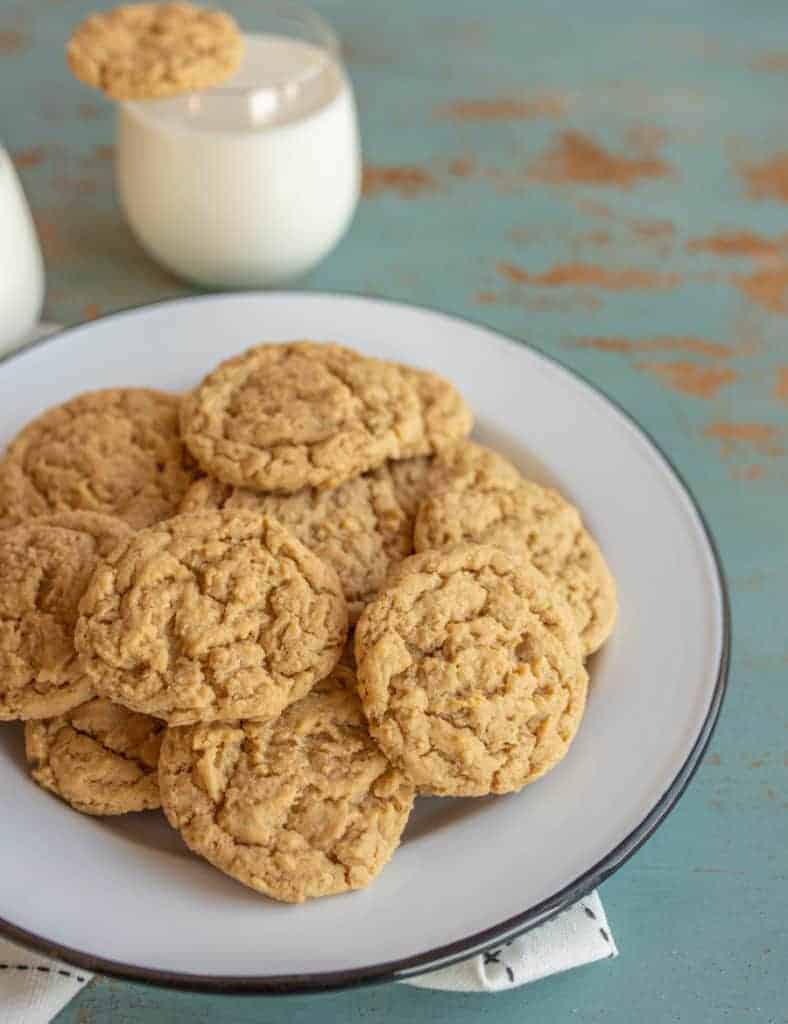 A white plate piled with peanut butter oatmeal cookies sits on a blue rustic table. In the background, two glasses of milk are visible, one with a cookie resting on top.
