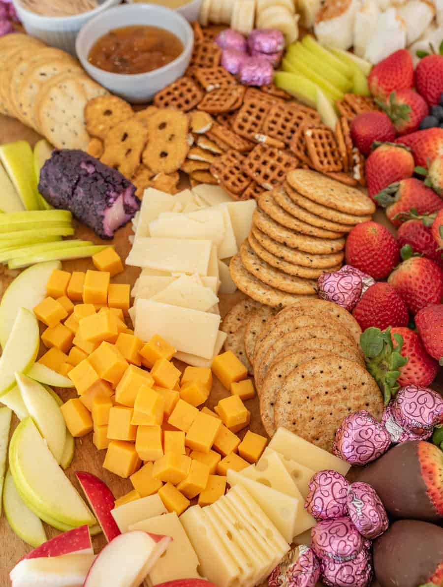 A close up of some of the elements on the chocolate and cheese board, including blueberry goat cheese, Swiss cheese, cubes of cheddar cheese, apple slices, strawberries, pretzel crisps, and crackers, garnished with heart-shaped chocolates. 