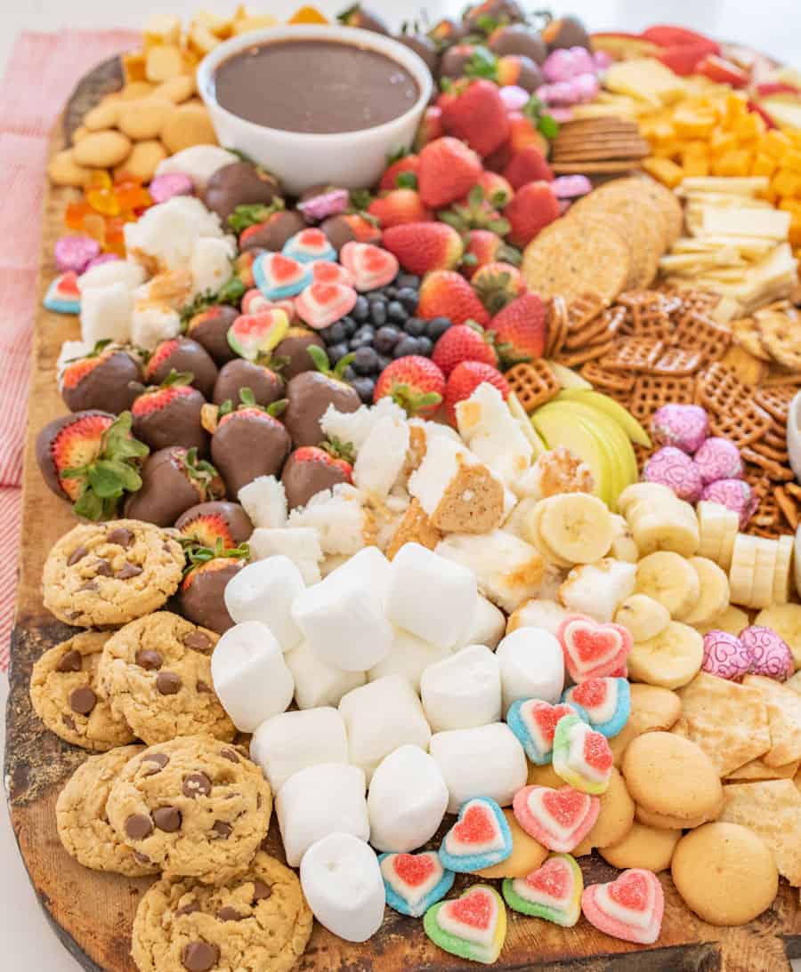 A large wooden board covered in cheeses, crackers, jam, mustard, toothpicks, chocolate fondue and things to dip in the fondue including fruit, chocolate chip cookies, vanilla wafers, banana slices, marshmallows, and garnished with heart-shaped gum drops and chocolates. 