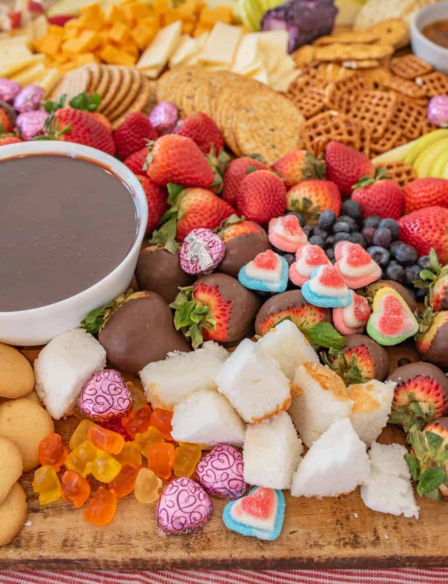 A close up of the chocolate fondue on the cheese board  with things to dip in the fondue including pretzels, apple slices, strawberries,  blueberries, and garnished with heart-shaped chocolates and gum drops. 