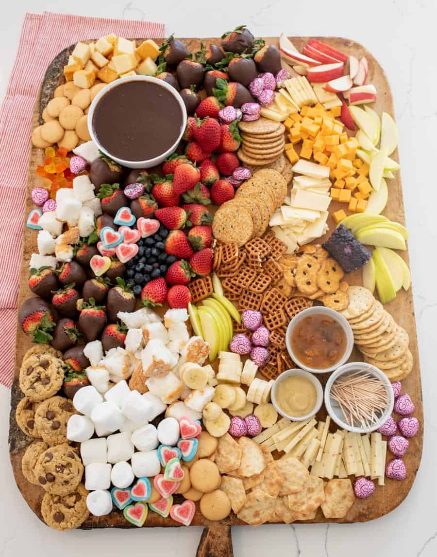 A large wooden board covered in cheeses, crackers, jam, mustard, toothpicks, chocolate fondue and things to dip in the fondue including fruit, cookies, marshmallows, and garnished with heart-shaped gum drops. 