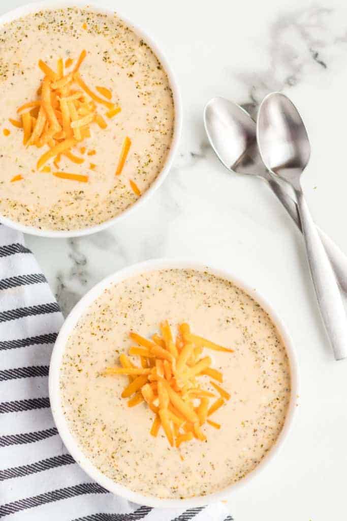 overhead shot of two bowls of cheesy broccoli soup topped with shredded cheese.