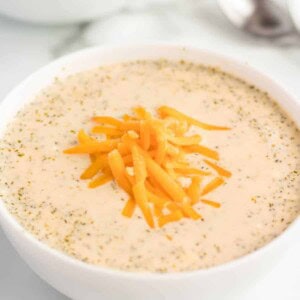 A white bowl filled with creamy crockpot broccoli and cheese soup, topped with shredded cheddar. A spoon and another bowl are visible in the blurred background.