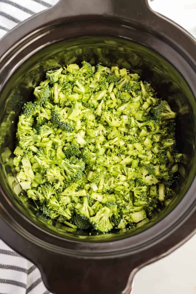 Chopped broccoli florets fill the inside of a black slow cooker, ready to be cooked. The photo is taken from above, showing the fresh green broccoli pieces.