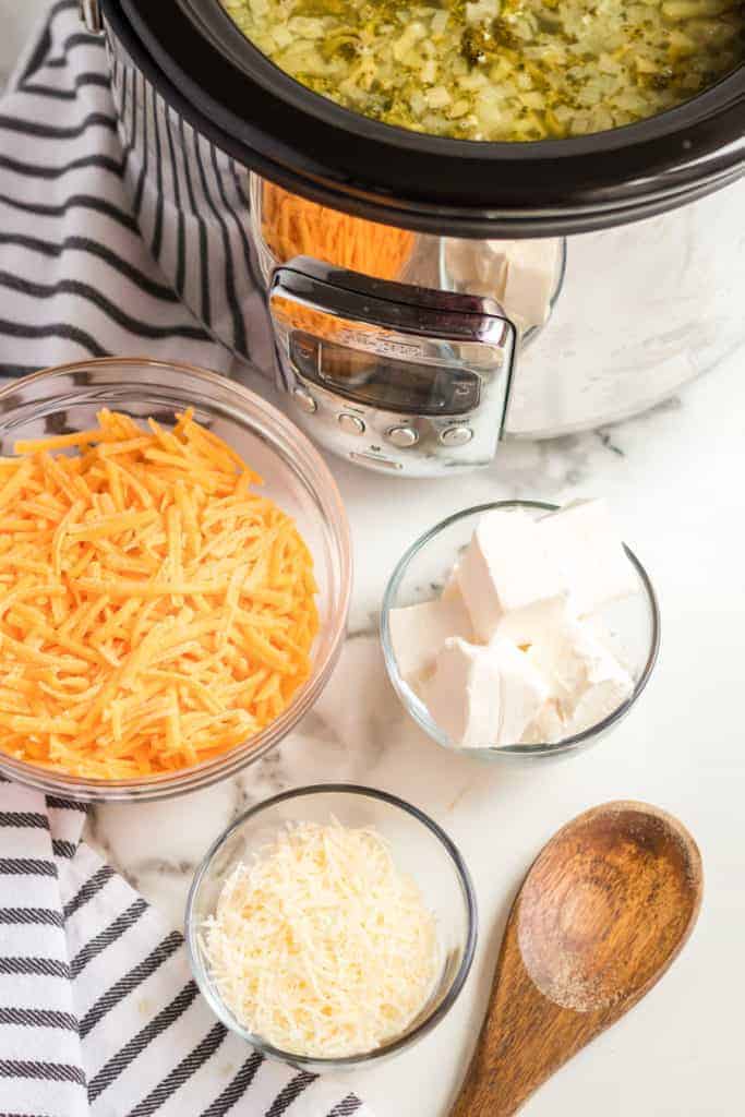 A slow cooker of Cheesy Broccoli Soup in the Crockpot sits on a marble counter next to a wooden spoon, shredded cheddar cheese, cubed cream cheese, shredded Parmesan, and a black-and-white striped towel.