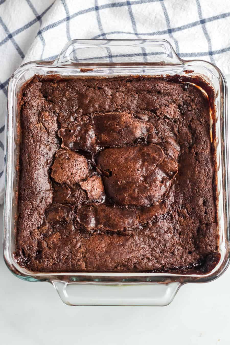 clear square pan with uncut chocolate brownie dessert in the pan sitting on a white and gray towel.