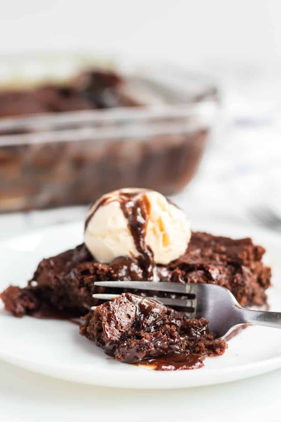 chocolate brownie dessert on a white plate with pale vanilla ice cream scoop on top and a drizzle of chocolate sauce with a fork in the brownie.