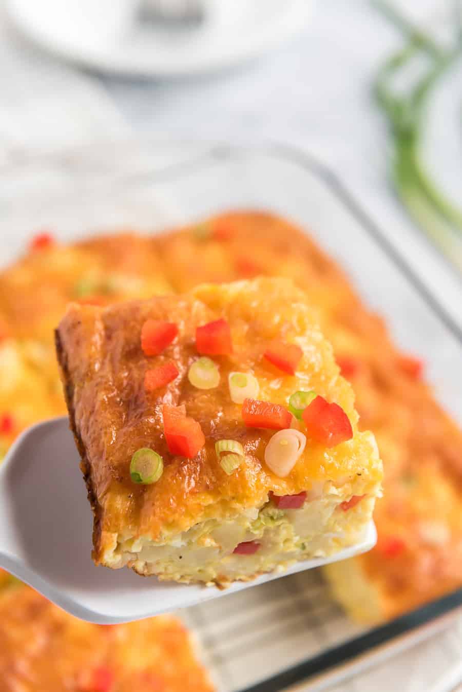 piece of egg and potato breakfast casserole on a spatula with little pieces of red pepper and green onions sprinkled on top and rest of casserole in pan in the background.
