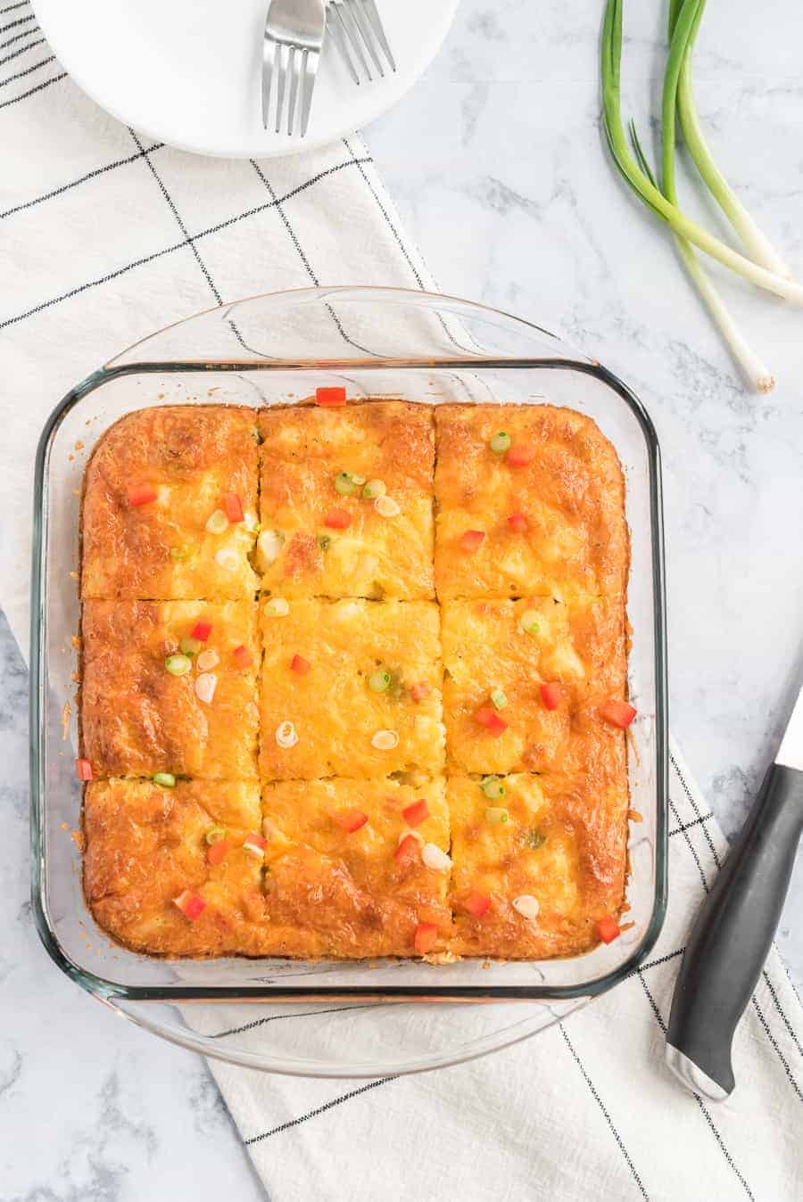 clear baking pan with egg and potato breakfast casserole with little pieces of red pepper and green onions sprinkled on top on white towel with black handled knife beside.