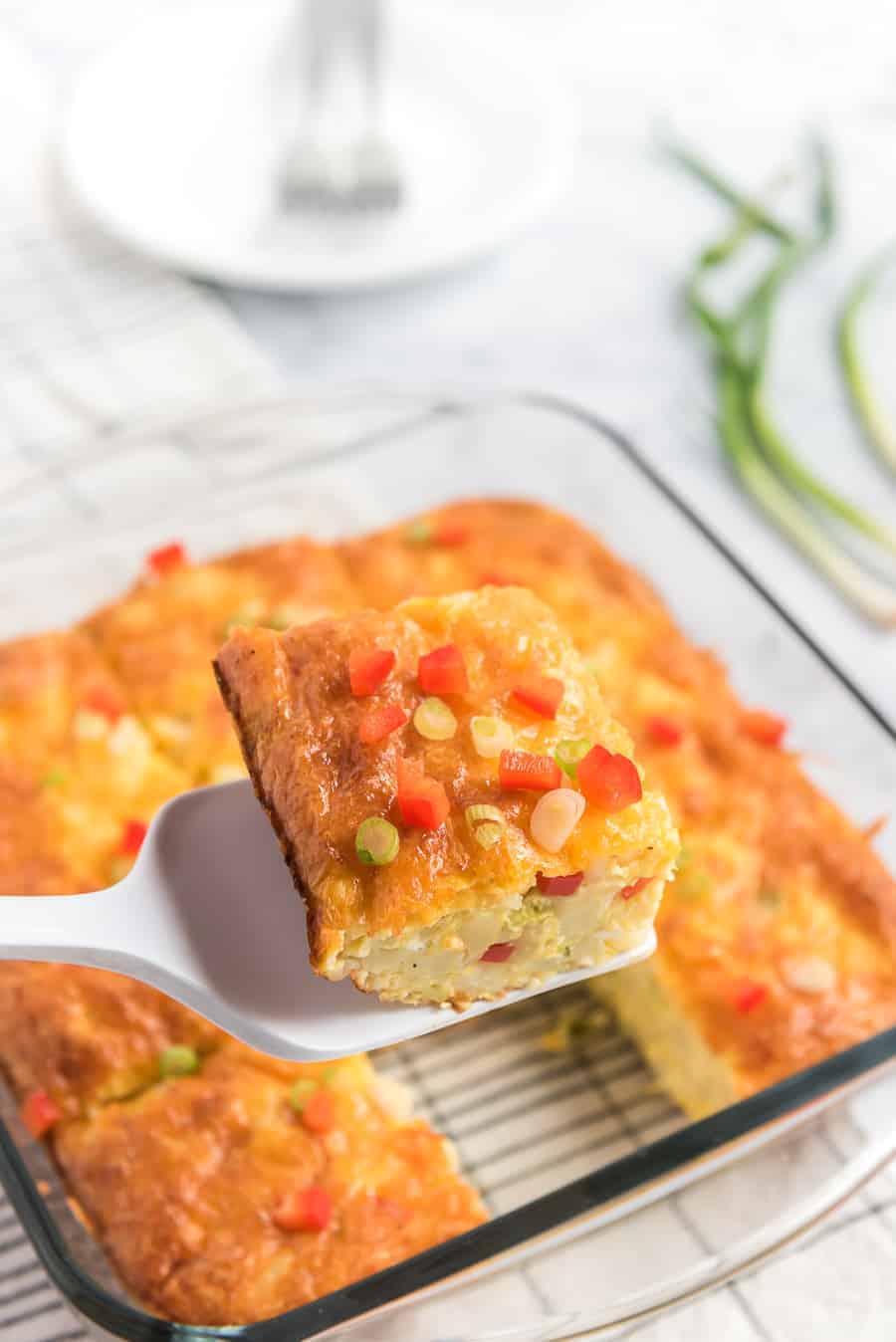 piece of egg and potato breakfast casserole on a spatula with little pieces of red pepper and green onions sprinkled on top and rest of casserole in pan in the background.