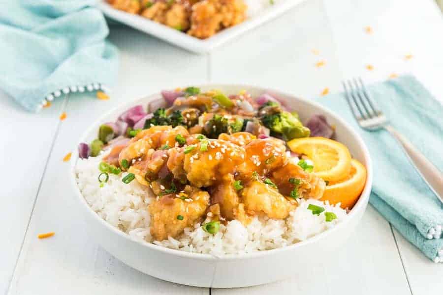 orange chicken on white rice with green broccoli, red onions, orange slices and green onions in a white bowl.