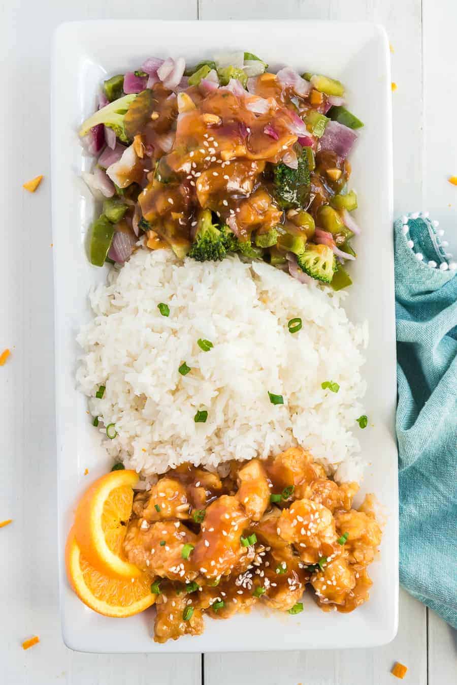 orange chicken on white rice with green broccoli, red onions, orange slices and green onions in a white bowl.