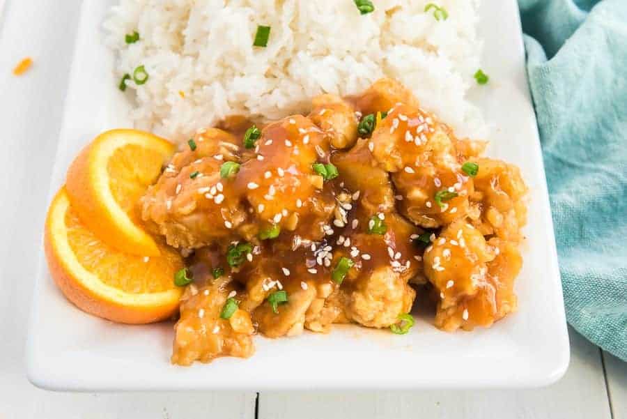 orange chicken on white rice with green broccoli, red onions, orange slices and green onions in a white bowl.