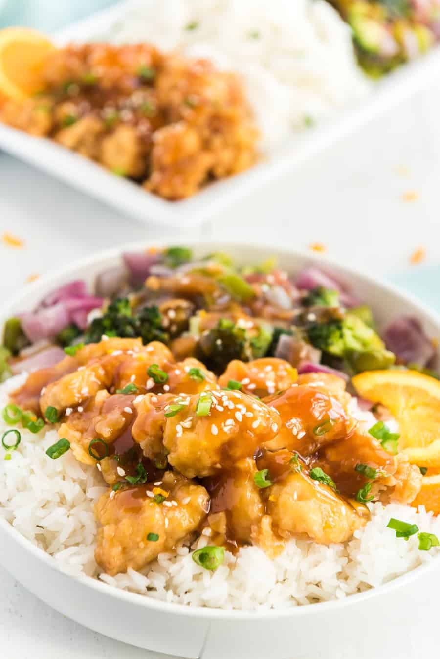 orange chicken on white rice with green broccoli, red onions, orange slices and green onions in a white bowl.