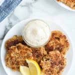 A white plate showcases four pan-fried salmon cakes, reminiscent of an old-fashioned salmon patties recipe, accompanied by a lemon wedge and a small glass bowl of creamy sauce. In the background, more salmon cakes are neatly arranged on the marble surface.