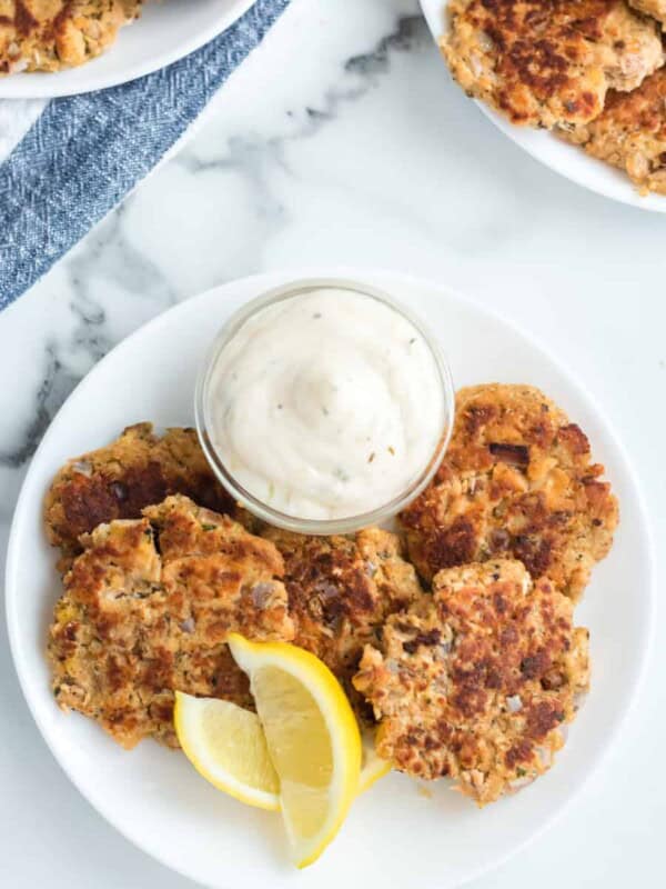 A white plate showcases four pan-fried salmon cakes, reminiscent of an old-fashioned salmon patties recipe, accompanied by a lemon wedge and a small glass bowl of creamy sauce. In the background, more salmon cakes are neatly arranged on the marble surface.