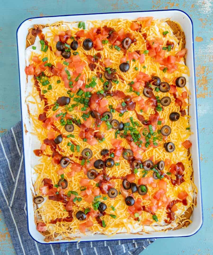 layered bean dip with cheddar cheese, tomatoes, black olives, and green onions on top in a white pan on a blue and white striped towel.