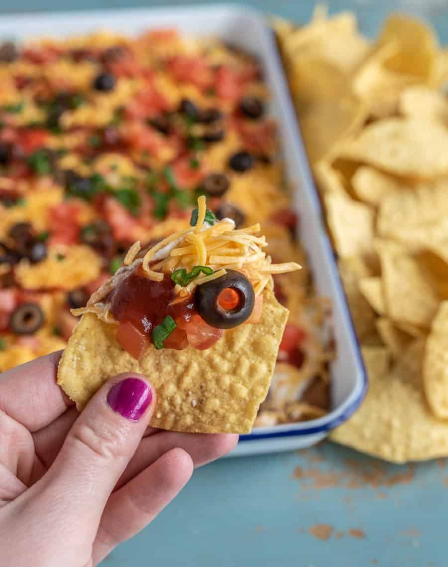 layered bean dip with cheddar cheese, tomatoes, black olives, and green onions on top with pile of tortilla chips beside it and a hand holding a chip with a scoop of dip.