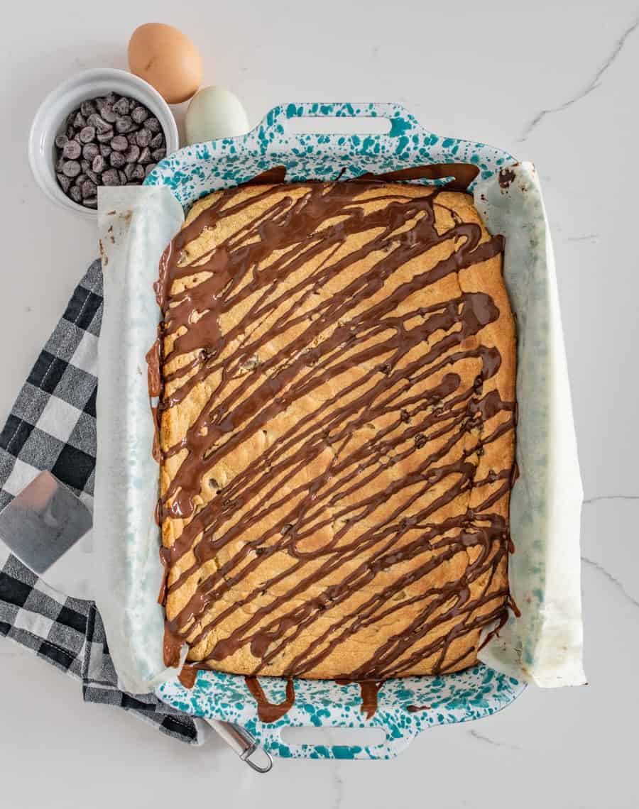 chocolate chip cookie bars in a blue and white pan with a bowl of chocolate chips and two eggs beside it on a black and white checkered towel.