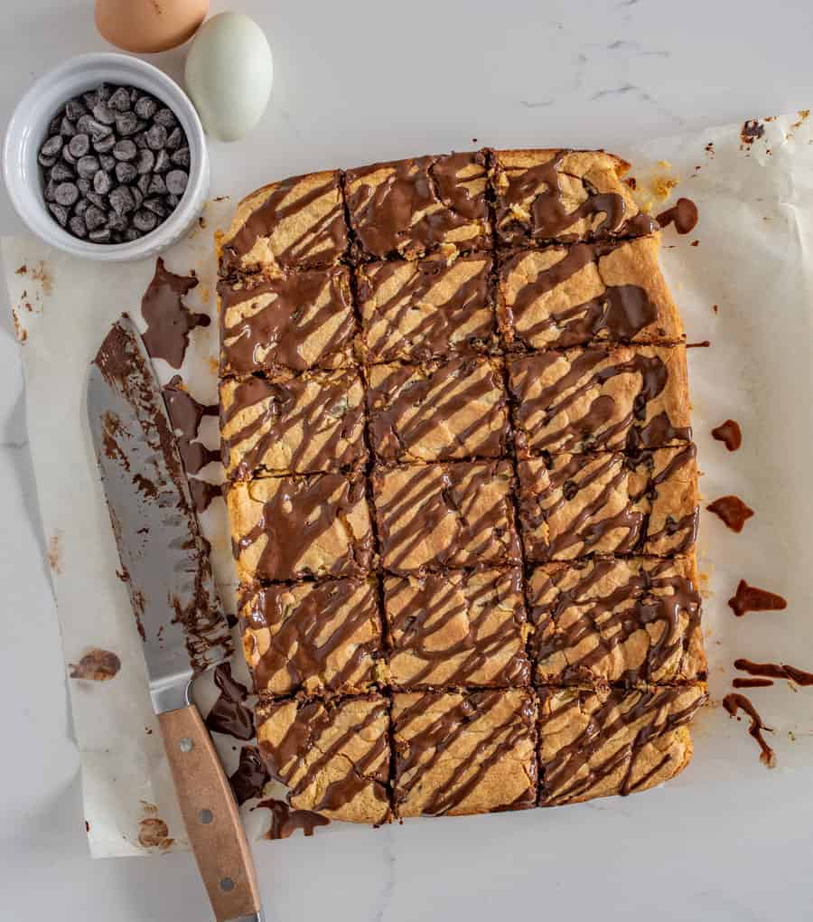 chocolate chip cookie bars on a white piece of parchment paper with a chocolate covered knife beside and a bowl of chocolate chips and two eggs beside it.