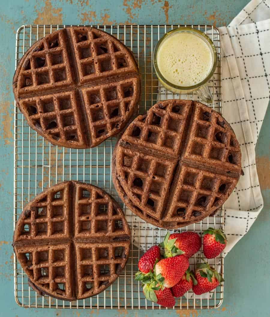 choclate waffle on a wire rack with whole strawberries and a cup of orange juice beside them on top of a white and black towel on a blue background.