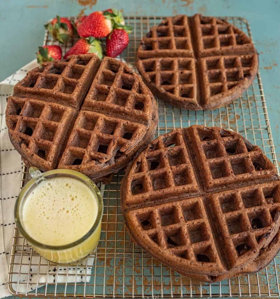 choclate waffle on a wire rack with whole strawberries and a cup of orange juice beside them on top of a white and black towel on a blue background.