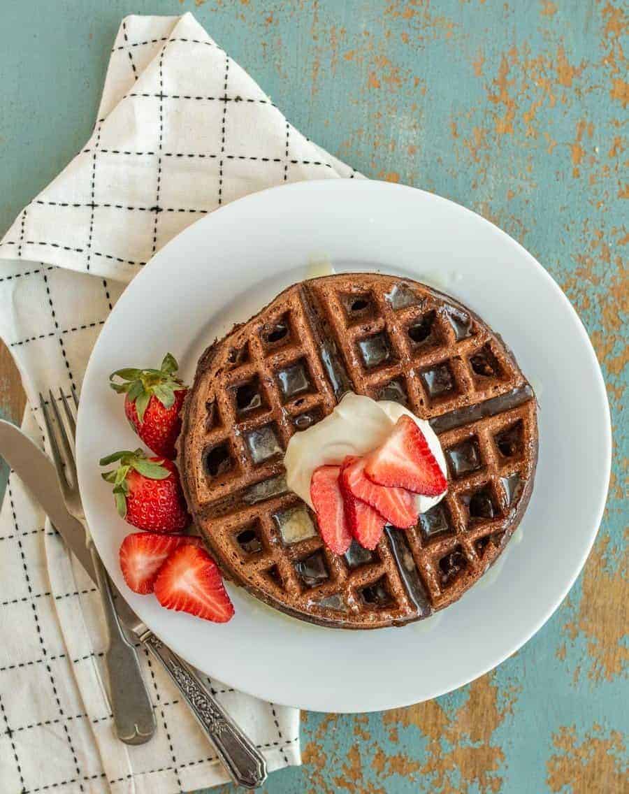 choclate waffle on a white plate with whole strawberries beside them and sliced strawberries, butter, and syrup on top.