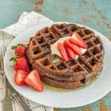choclate waffle on a white plate with whole strawberries beside them and sliced strawberries, butter, and syrup on top.