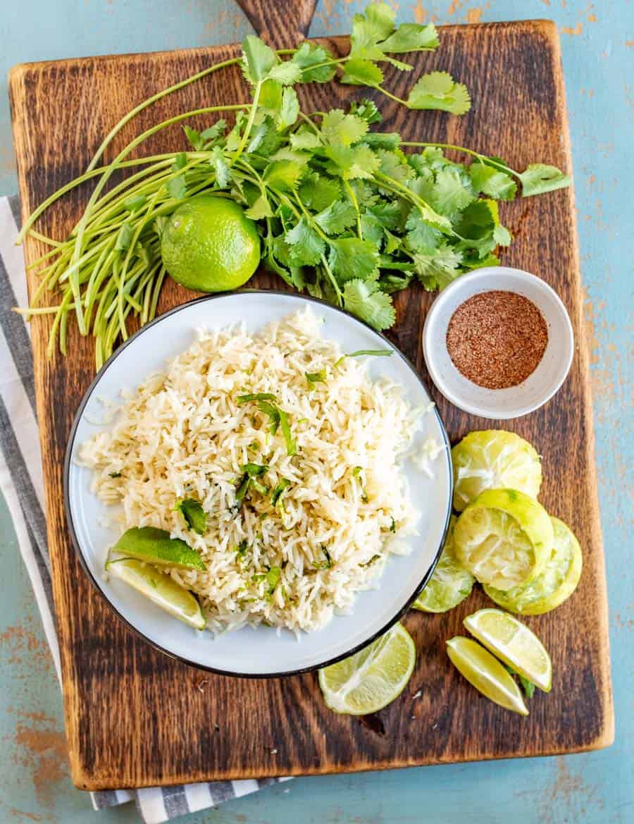 a plate of cilantro lime rice topped with fresh cilantro and lime wedges. Plate is surrounded by cilantro, limes, and spices.