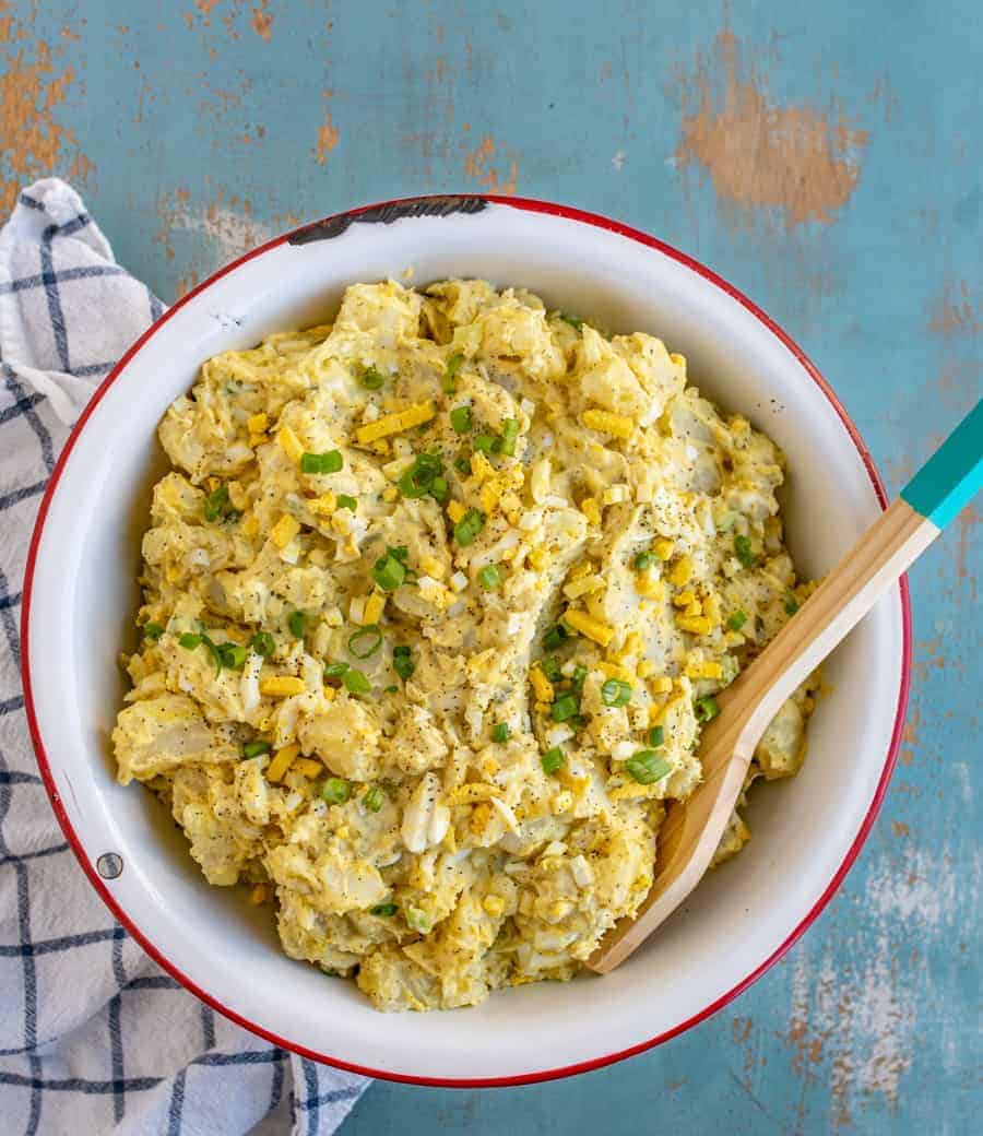 white bowl with red rim filled with yellow-ish potato salad with chopped eggs and green onions mixed in with a wooden spoon sticking out and sitting on a blue and white towel.