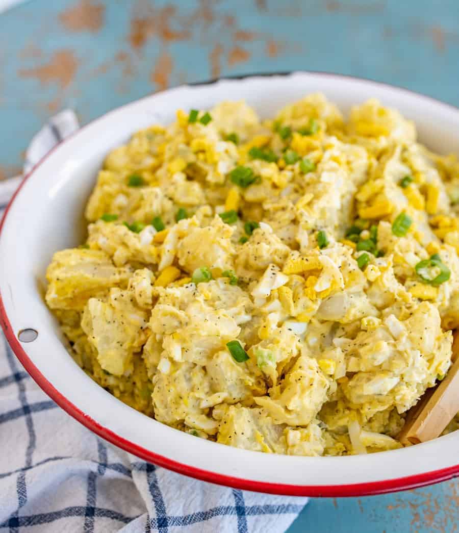 white bowl with red rim filled with yellow-ish potato salad with chopped eggs and green onions mixed in with a wooden spoon sticking out and sitting on a blue and white towel.