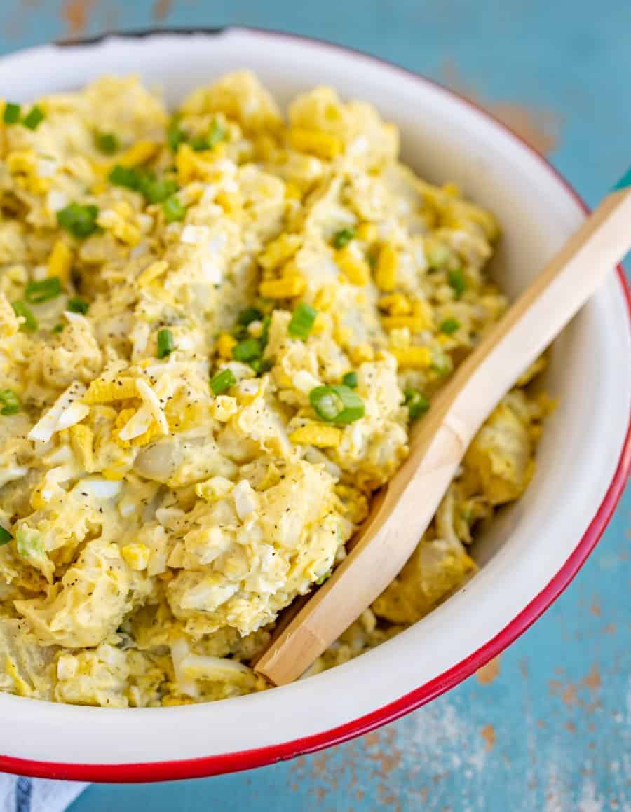 classic potato salad with a yellow tint with chopped eggs and green onions mixed in set in a white bowl with a red rim and a wooden spoon sticking out.