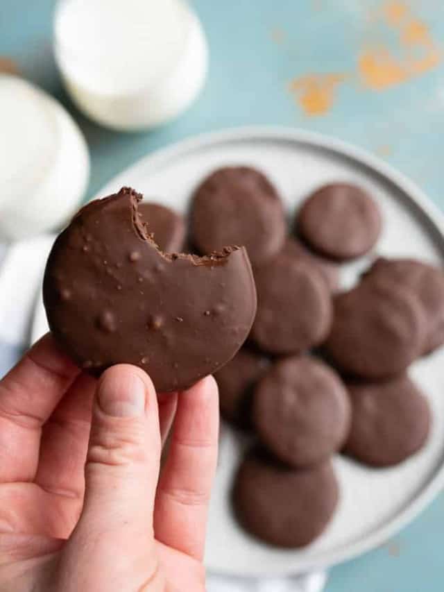 round chocolate cookies on white plate with two glasses of white milk and light blue background and one cookie held in hand.