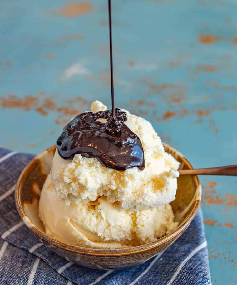 homemade hot fudge sauce poured over vanilla ice cream in a bowl with a spoon on a blue and white striped towel.