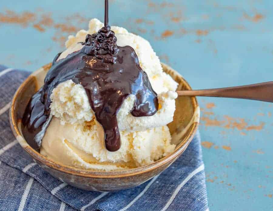 homemade hot fudge sauce poured over vanilla ice cream in a bowl with a spoon on a blue and white striped towel.