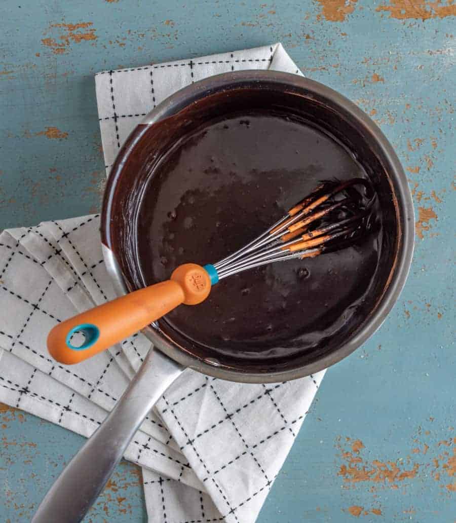 chocolate sauce in a metal pan on a black and white towel with a metal whisk.