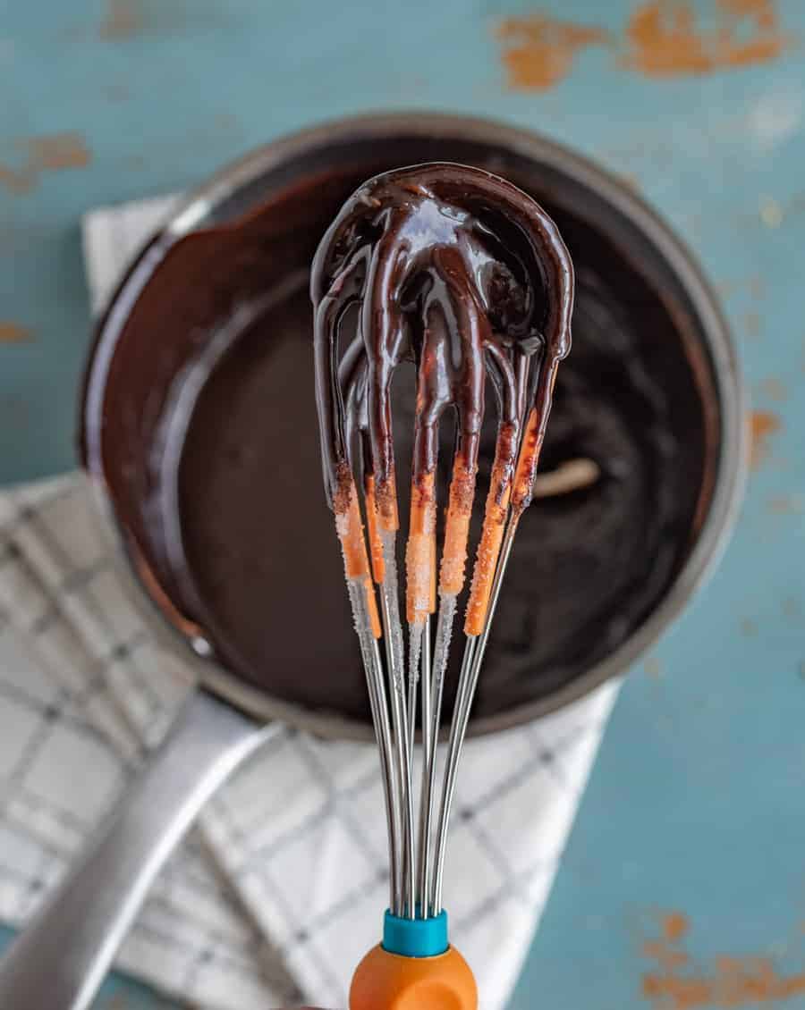 chocolate sauce in a metal pan on a black and white towel with a metal whisk.