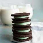 stack of four homemade mint oreo cookies with dark chocolate outside and mint green inside stacked on a white towel with two clear glasses of white milk behind.