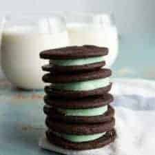 stack of four homemade mint oreo cookies with dark chocolate outside and mint green inside stacked on a white towel with two clear glasses of white milk behind.