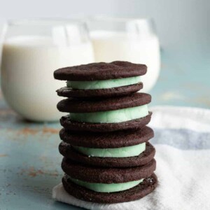 stack of four homemade mint oreo cookies with dark chocolate outside and mint green inside stacked on a white towel with two clear glasses of white milk behind.