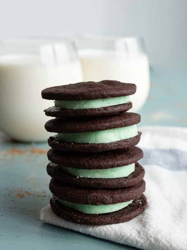 stack of four homemade mint oreo cookies with dark chocolate outside and mint green inside stacked on a white towel with two clear glasses of white milk behind.