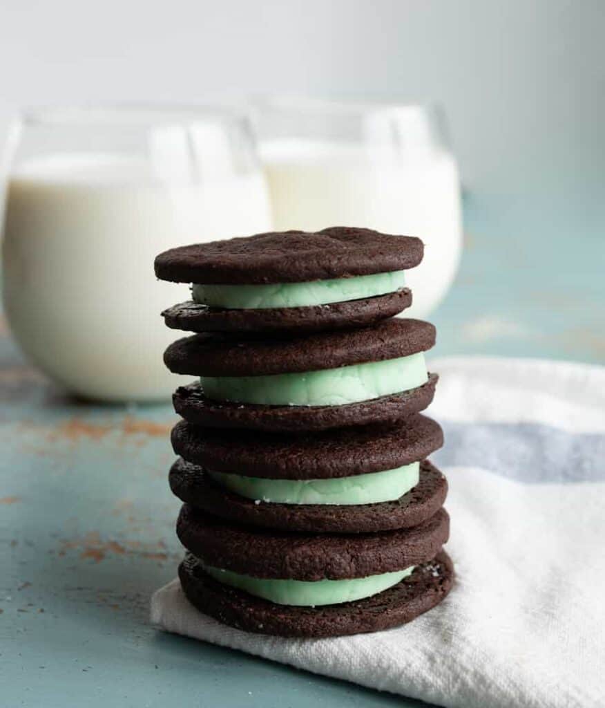 stack of four homemade mint oreo cookies with dark chocolate outside and mint green inside stacked on a white towel with two clear glasses of white milk behind.