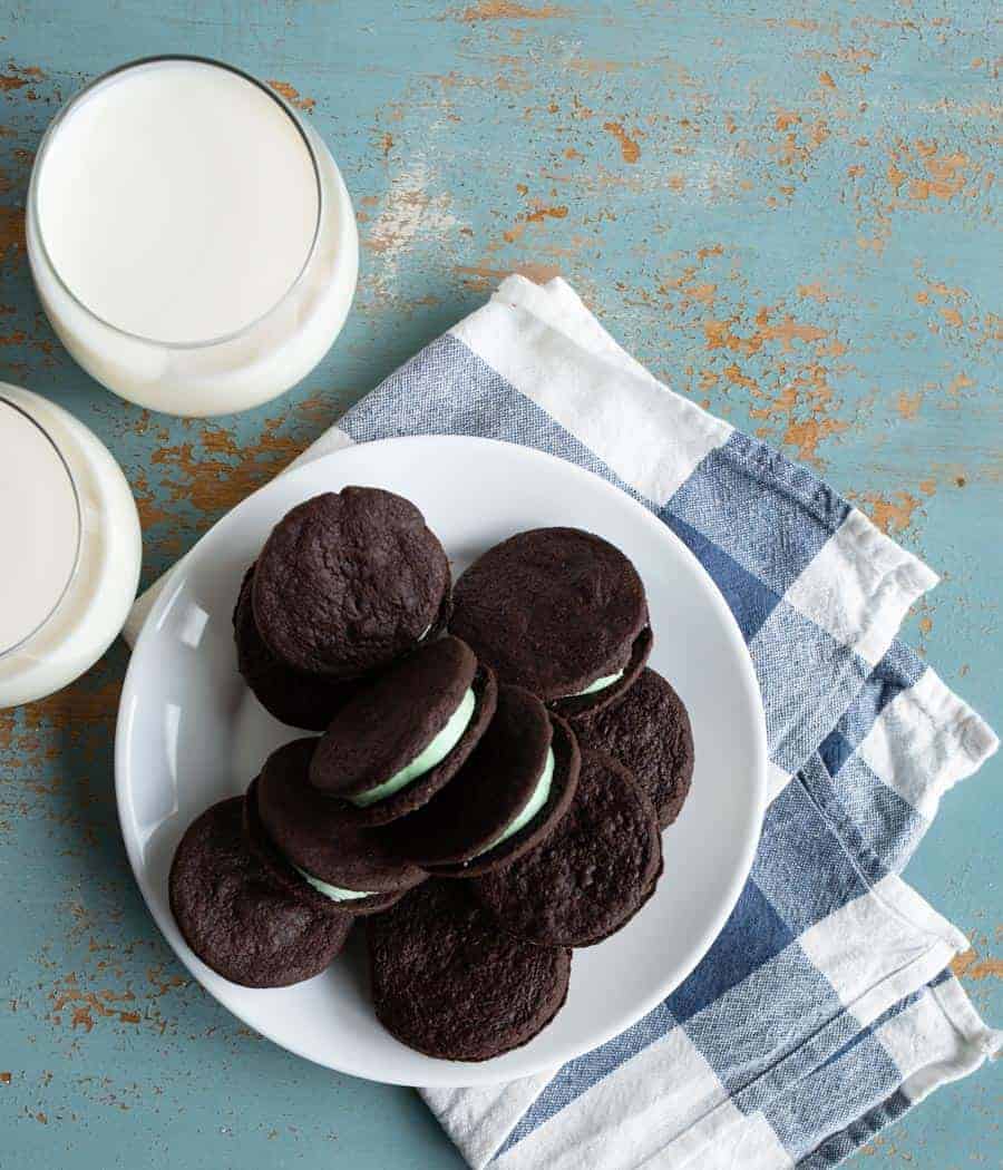 pile of homemade mint oreo cookies with dark chocolate outside and mint green inside stacked on a white plate on a blue and white towel on a blue background with two clear glasses of white milk beside them.