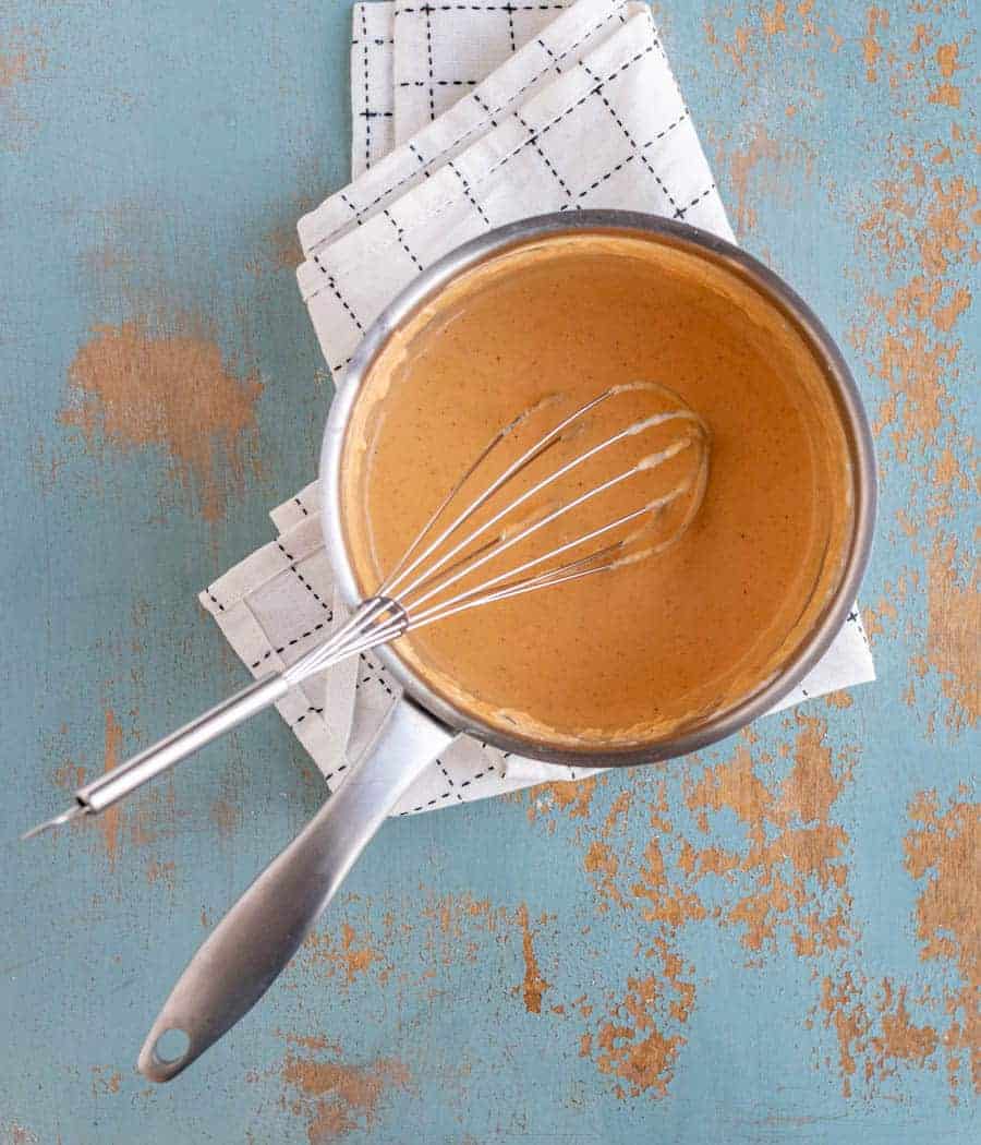 orange colored cheese sauce in metal pan with metal whisk on top of black and white towel on blue background.