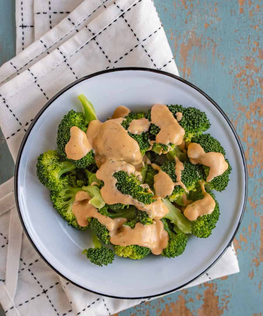 green broccoli florets on a white plate with cheese sauce on top sitting on black and white towel on a blue background.