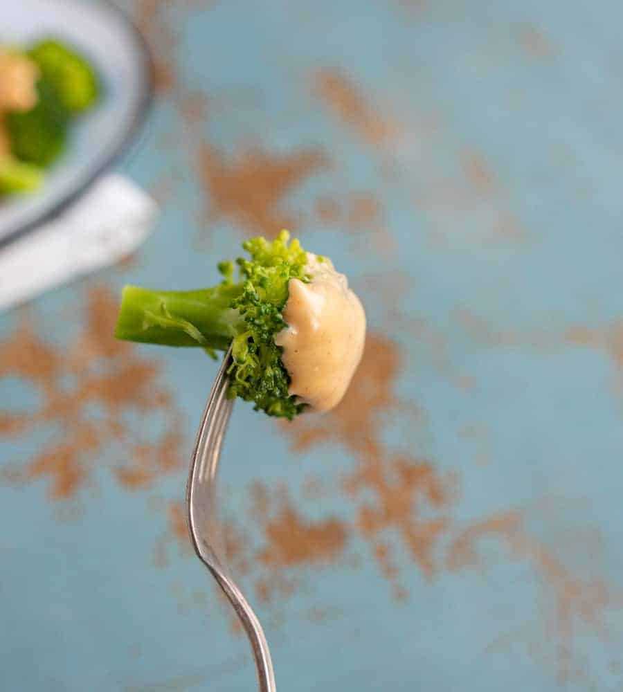green broccoli florets on a fork with cheese sauce on top on a blue background with blurry edge of plate with broccoli in the background.