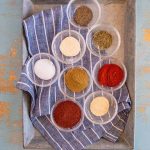 small clear glass bowls with different colored spices in each set in metal pan on top of blue and white striped towel.