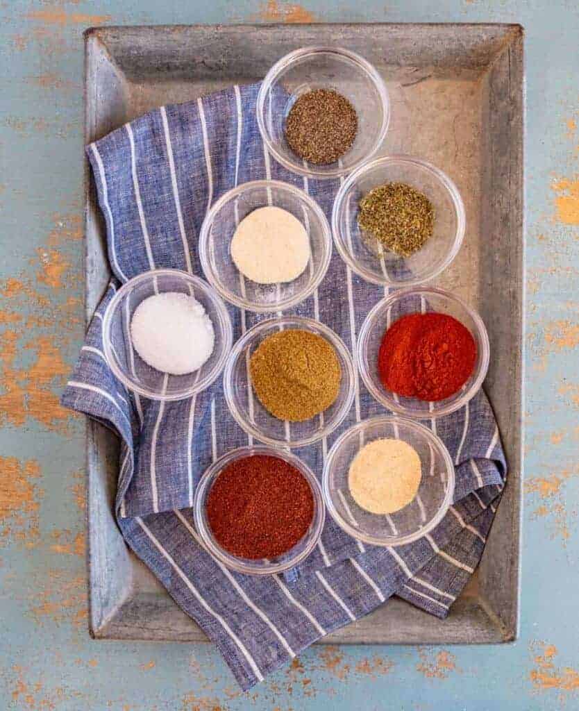 small clear glass bowls with different colored spices in each set in metal pan on top of blue and white striped towel.